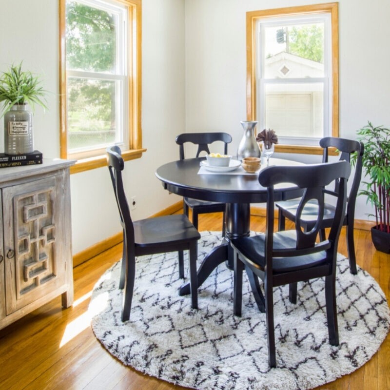 A room scene of a dining area with golden brown hardwood flooring and a round white area rug under a black dining table and chairs