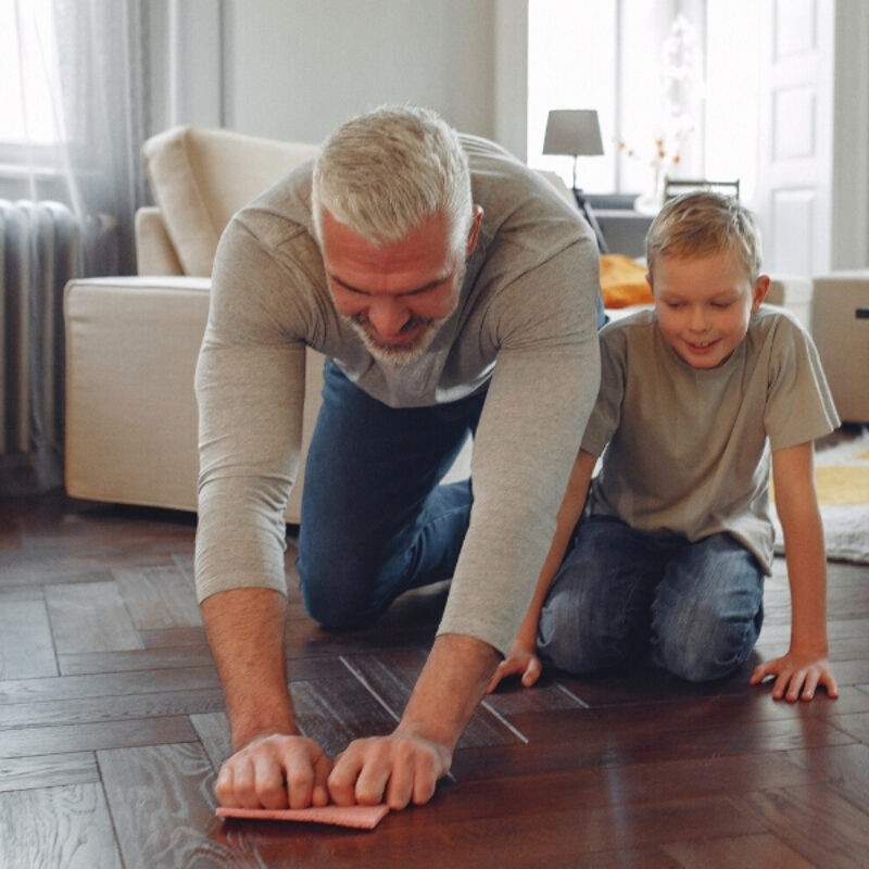 An image of a father and son kneeling on a herringbone floor with a cleaning rag in hand