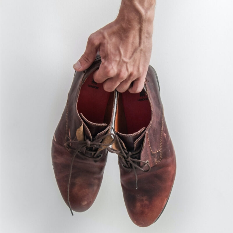 An image of a mans hand holding a pair of leather shoes in front of a white wall