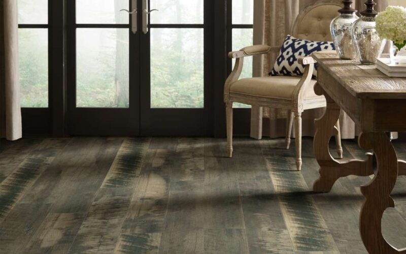 A room scene with a reclaimed wood look floor in a gray washed color. There are large black double doors in the background of the image looking out into a foggy woodsy scene. The furniture in the room is traditional and light in color.