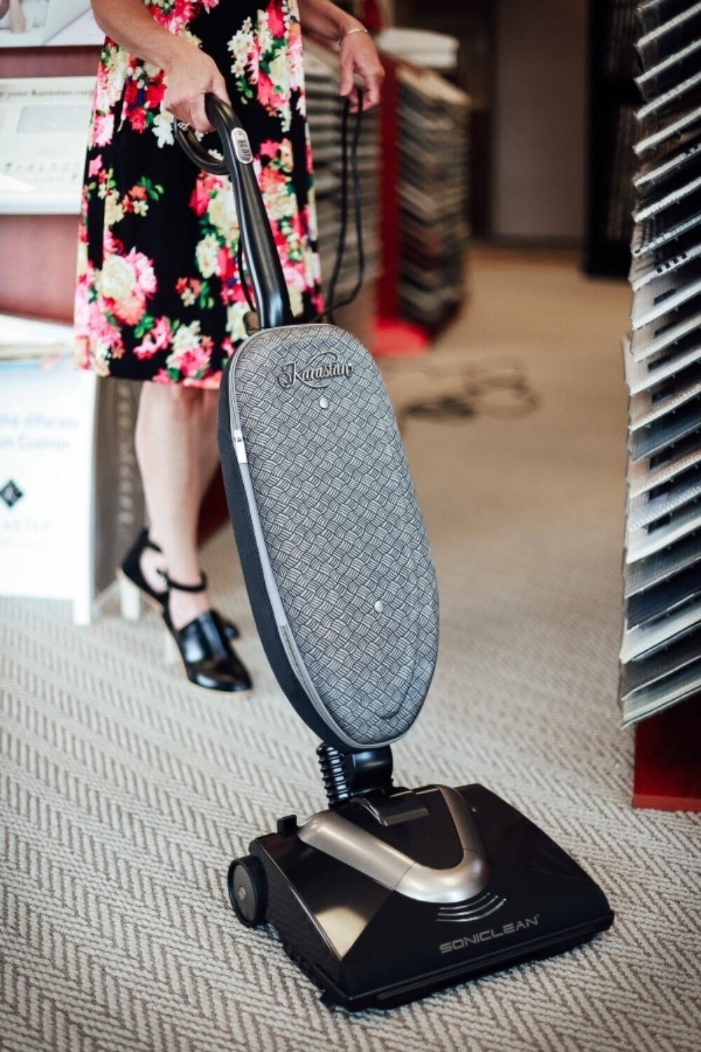 An image of a woman in a floral dress using the Karastan Soniclean vacuum on a woven look carpet. There are carpet display stands behind her.